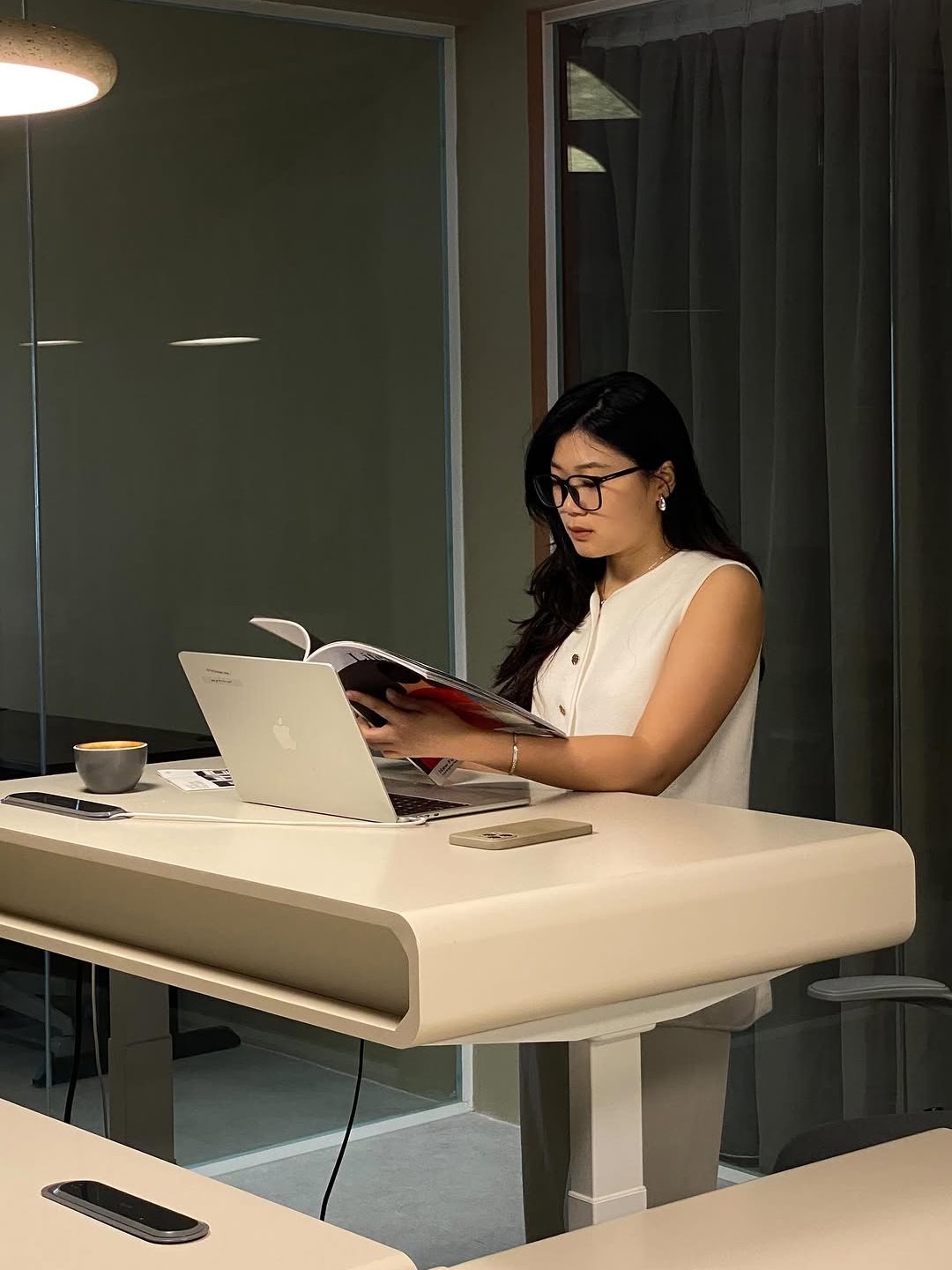 A person reading a magazine while working at a height-adjustable desk in a modern coworking space, with a laptop and coffee cup nearby.