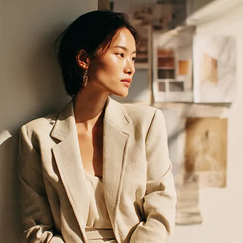 A focused woman in a cream oversized blazer leaning against a wall covered in design mood board references, bathed in warm natural light
