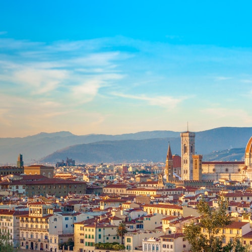 Panoramic view of Florence with its historic buildings and iconic cathedrals, under a clear blue sky with distant mountains.