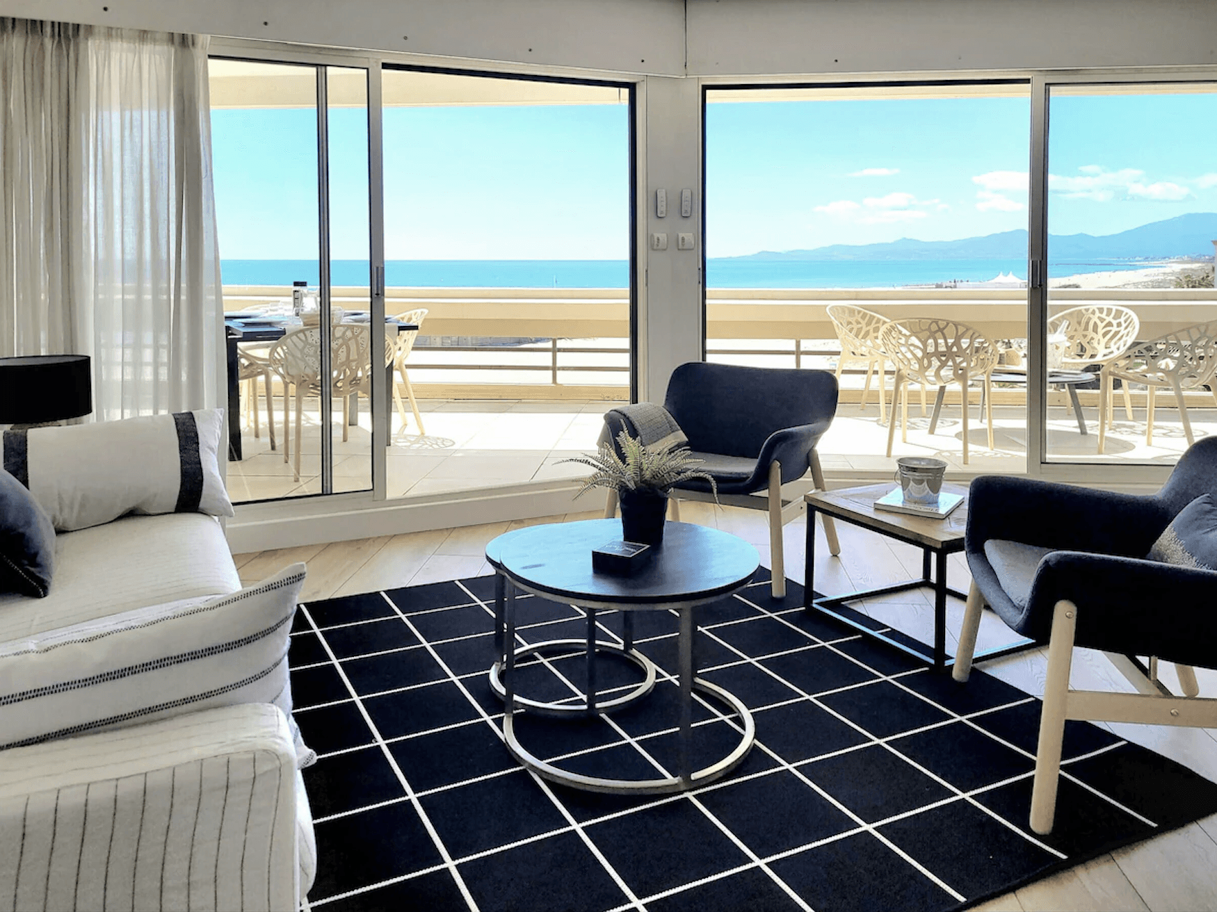 Two-story white house with a wraparound balcony overlooks a clear blue swimming pool. Palm trees and potted plants add a tropical vibe under a sunny sky.