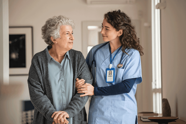Caregiver in scrubs supporting an elderly woman while walking indoors.