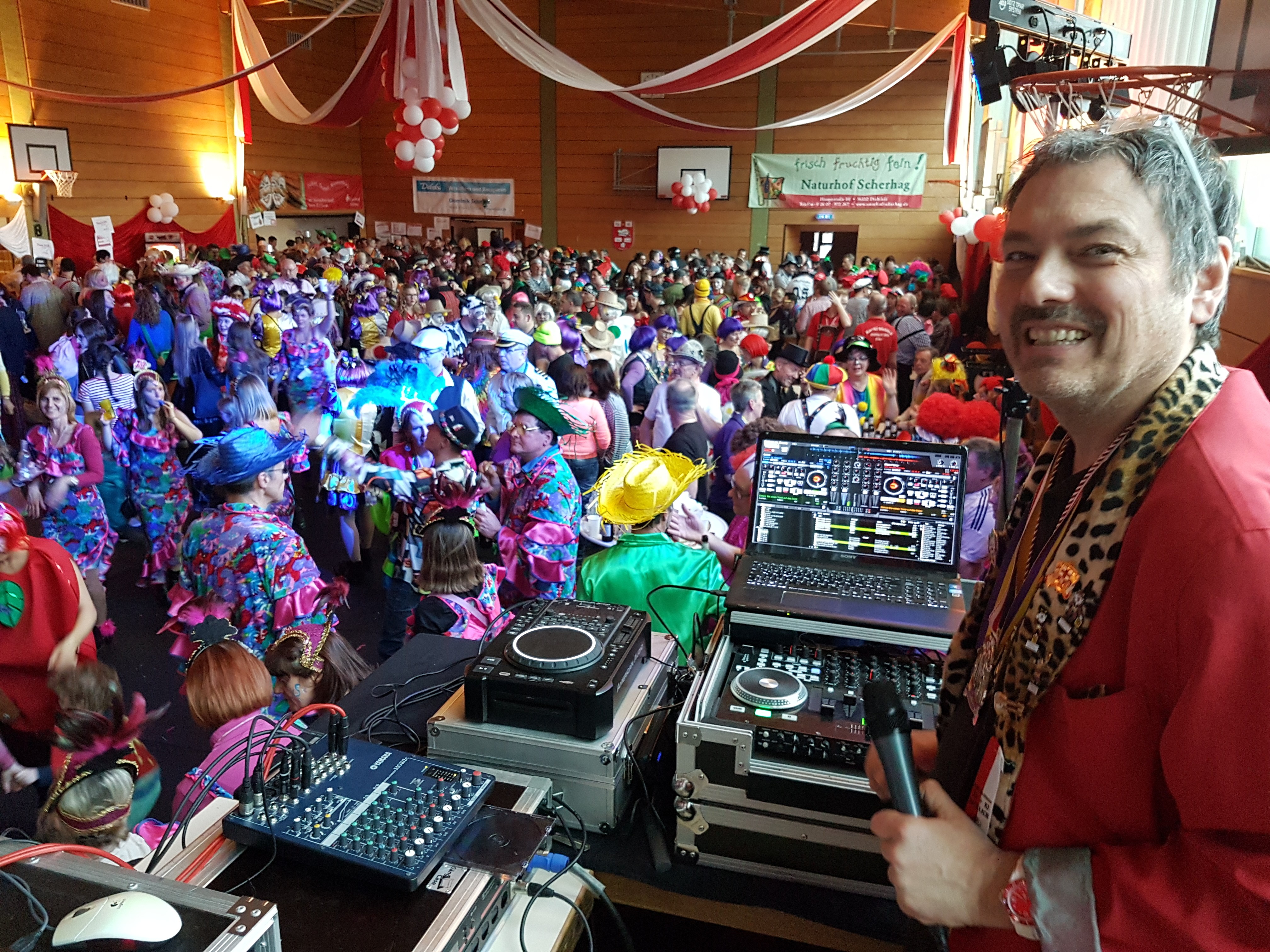 DJ lachend auf der Bühne vor Mischpult mit Mikrofon in der Hand. Dahinter eine mit Menschen gefüllte Halle. Menschen sind verkleidet, tanzen und haben gute Stimmung. DJ macht Musik und moderiert.