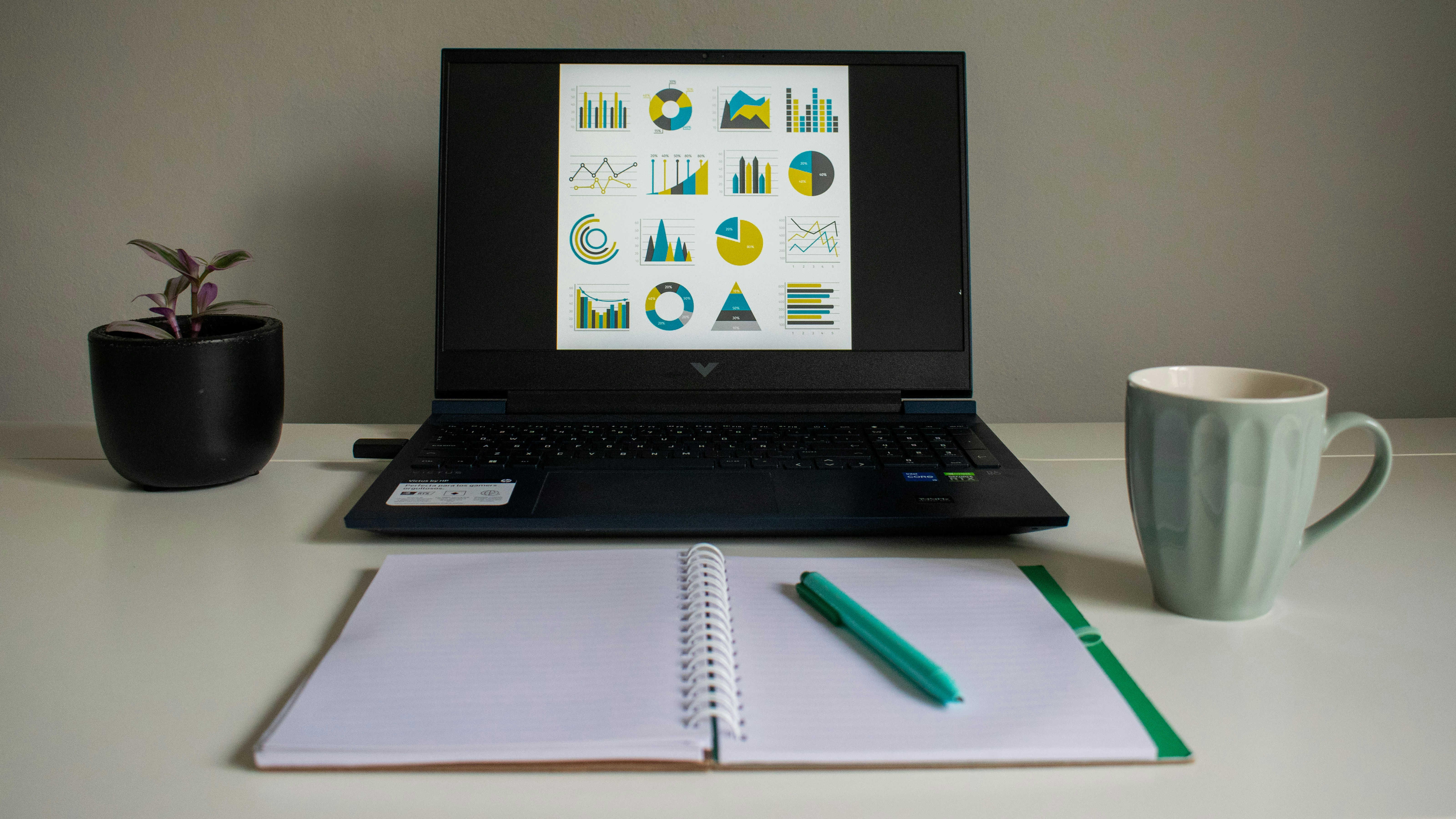 Laptop displaying charts next to notebook and mug.