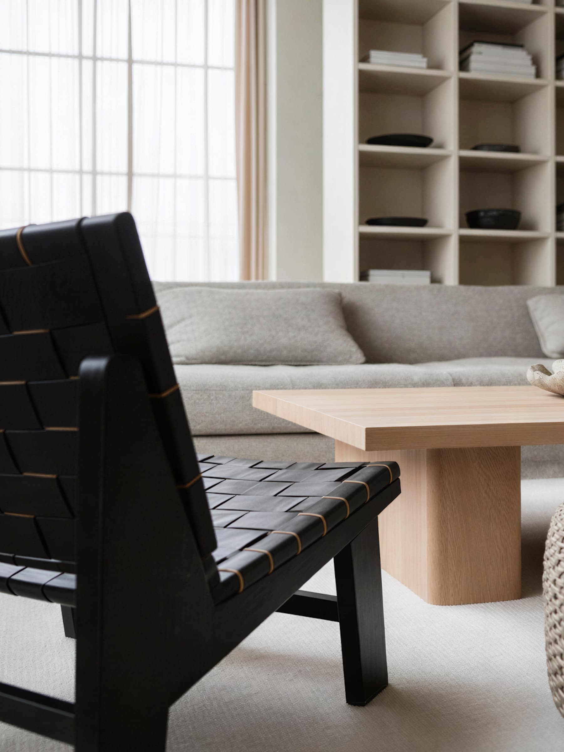 A modern living room features a black woven chair, a light wood coffee table, and a plush gray sofa set against a backdrop of minimalist shelves and sheer floor-to-ceiling curtains.