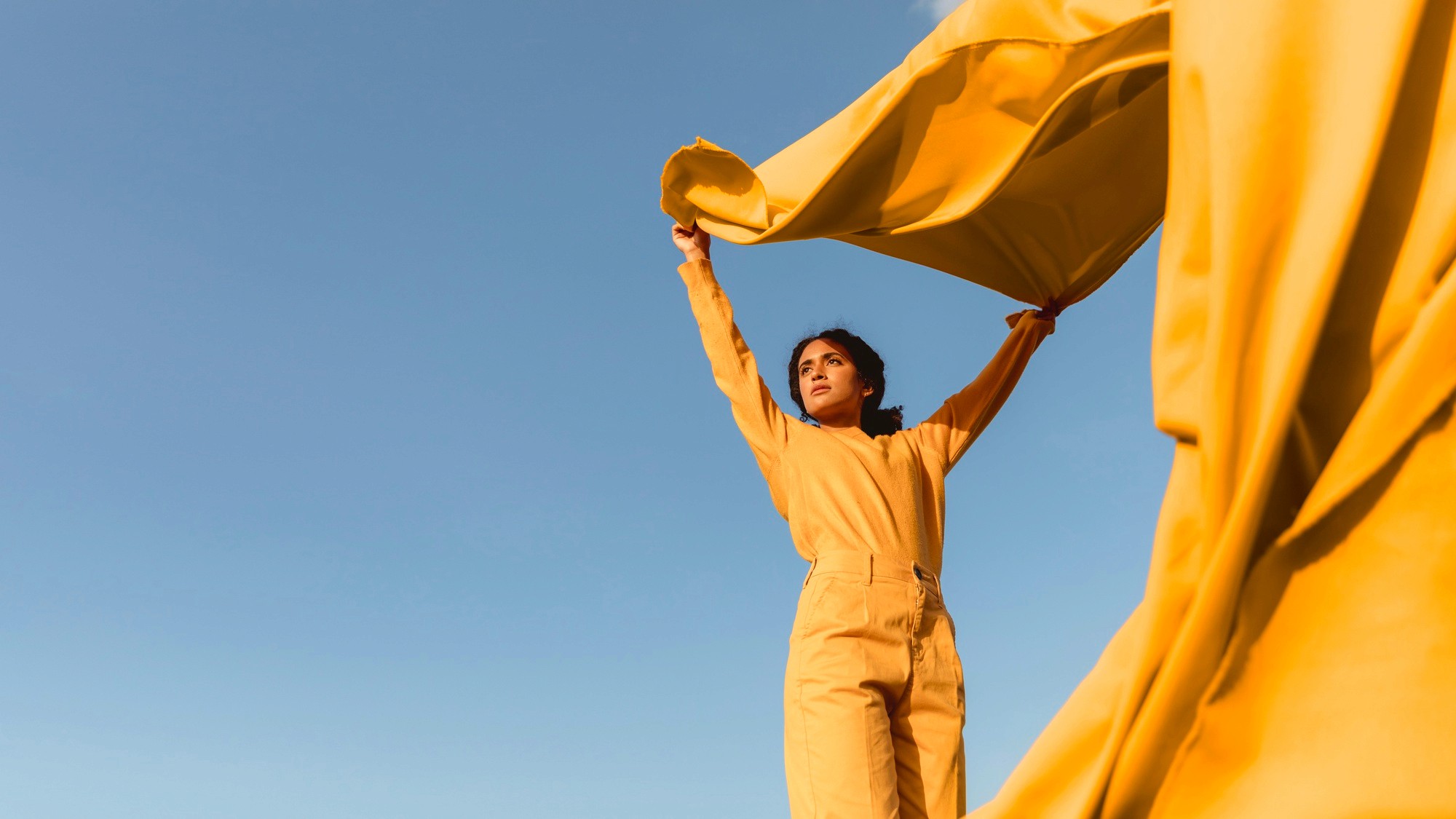 A person in yellow clothing holds up a large piece of yellow fabric against a clear blue sky.