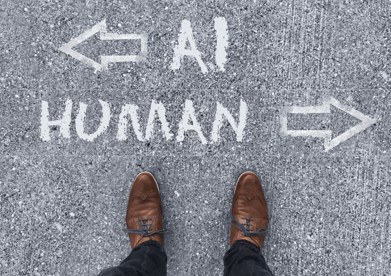Looking down at a pair of man's feet in brown dress shoes. In front of the shoes are the words "AI" with an arrow pointing left and "Human" with an arrow pointing right.