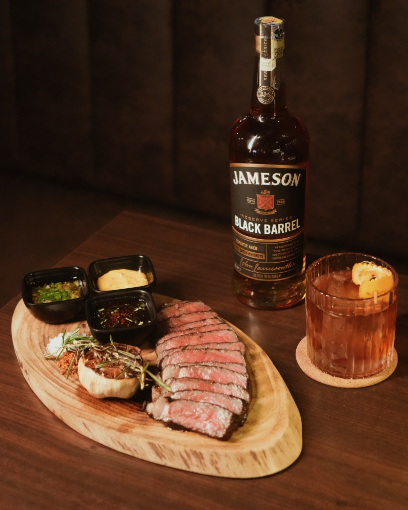 A wooden platter featuring sliced steak served with various dipping sauces, alongside a bottle of Jameson Black Barrel whiskey and a cocktail in a glass with an orange garnish.