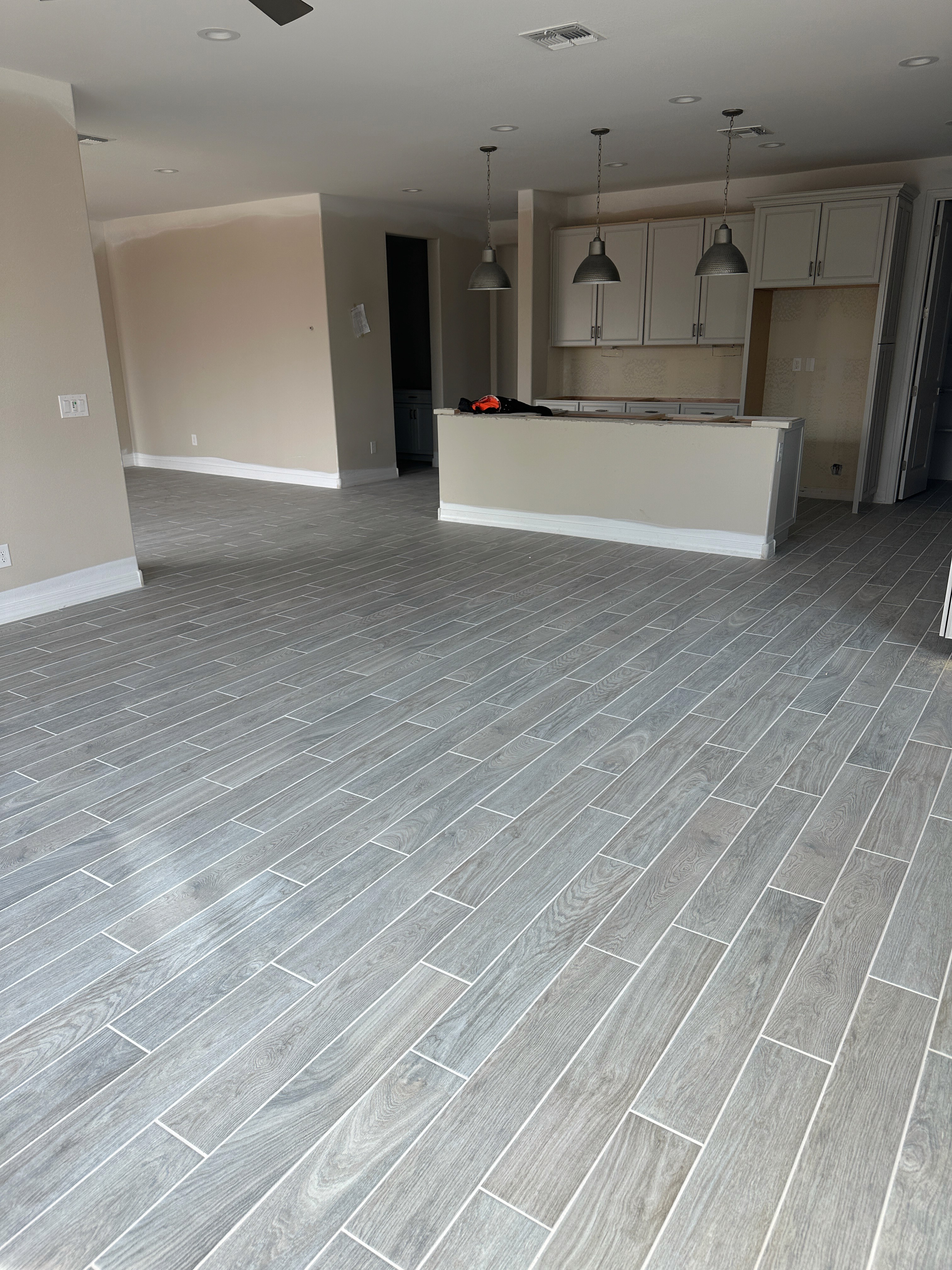 Modern open-plan living area with gray wood-look tile flooring, kitchen island, and pendant lights in a new home under construction.