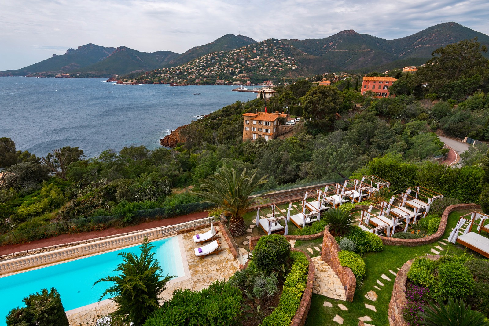 A coastal resort with a turquoise swimming pool and white lounge chairs overlooking a bay with Mediterranean villas.