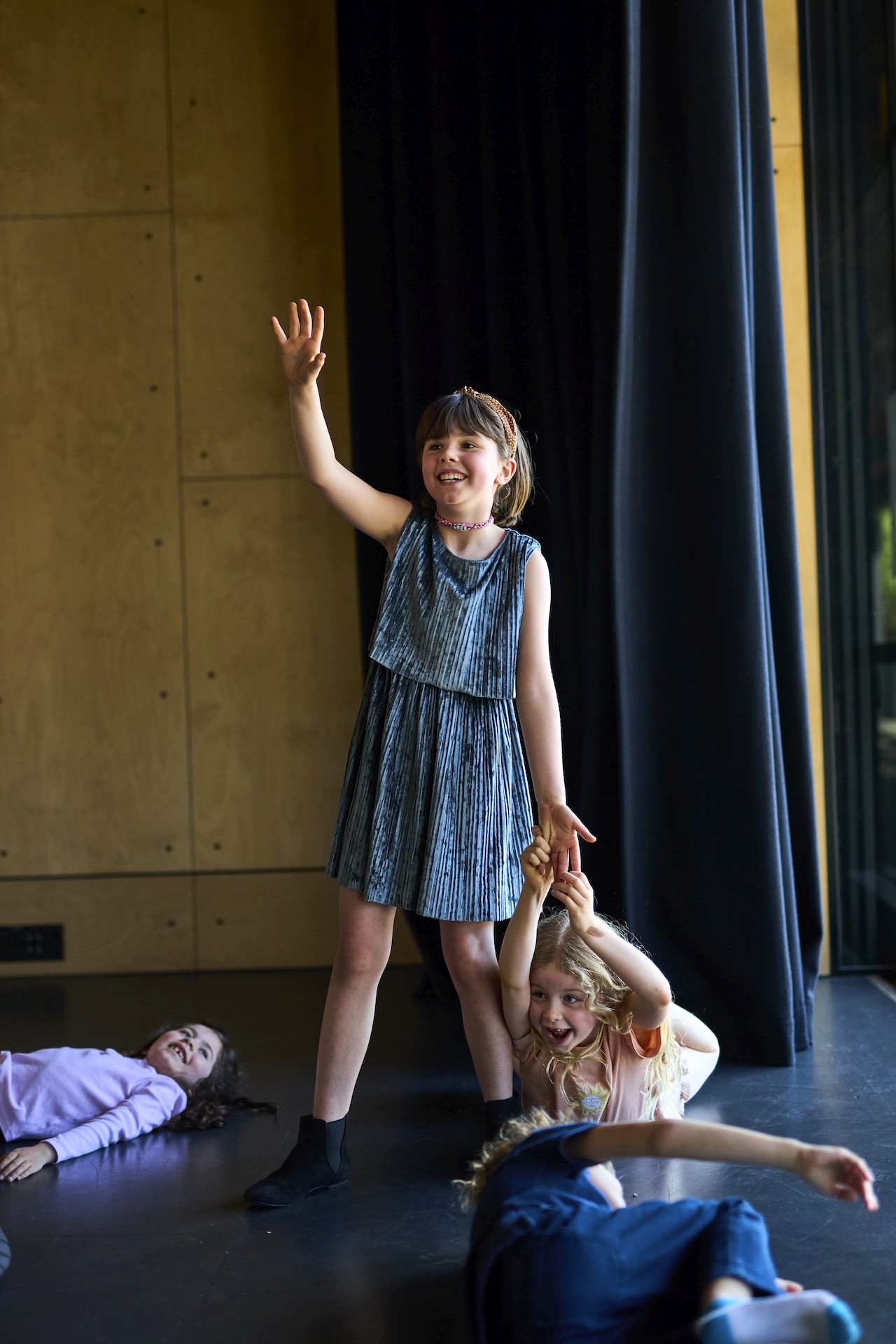 Three young performers stand on stage rehearsing a scene, watched by other students seated in the foreground.