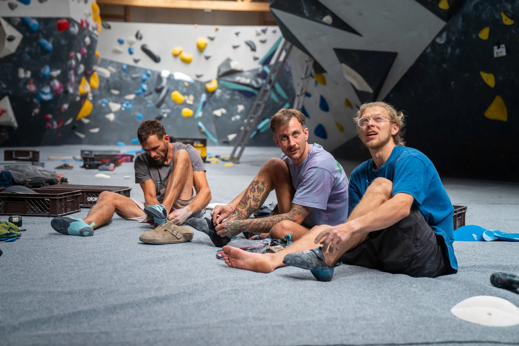 Three people sit on a climbing gym floor, smiling and surrounded by climbing gear and colorful climbing walls.