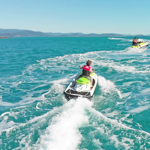 Two people on jet skis ride across clear, turquoise waters, leaving white wakes in their path under a bright blue sky.