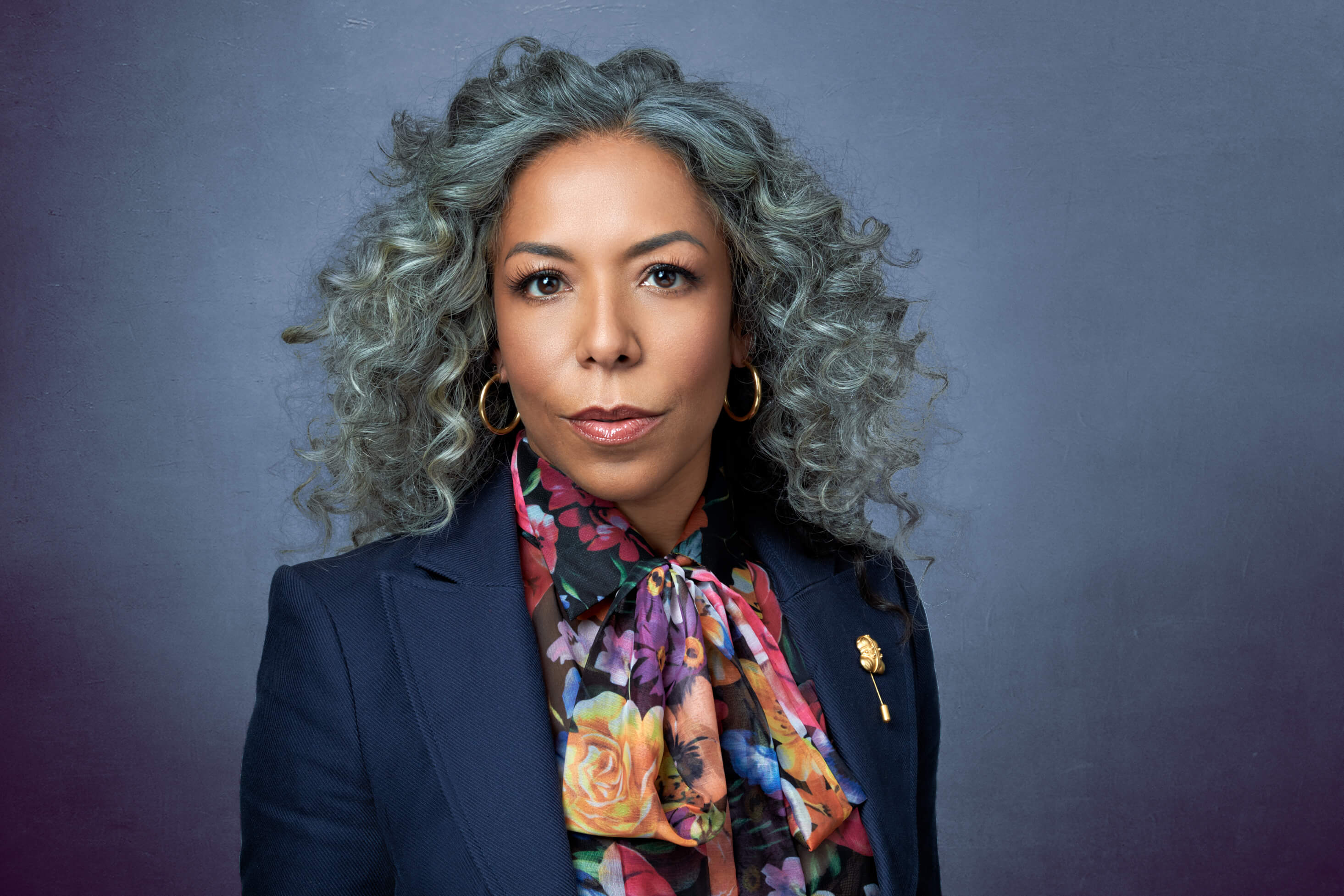 Studio headshot of mature Latina woman with silver curly hair in navy blazer and floral scarf