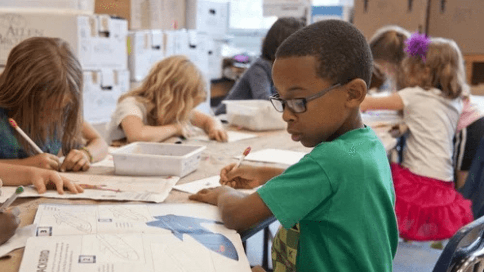Elementary school students sit at a classroom table working on worksheets and drawings, with one child in glasses focused on writing while others work nearby.