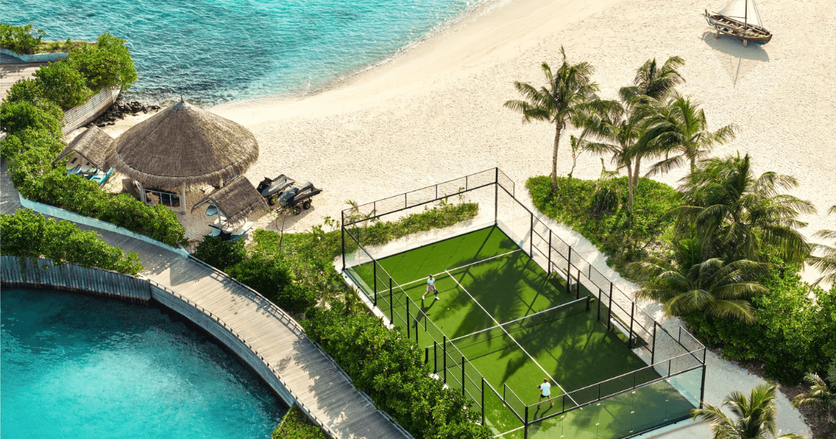 A padel court beside turquoise ocean water and white sandy beach at a tropical island resort.
