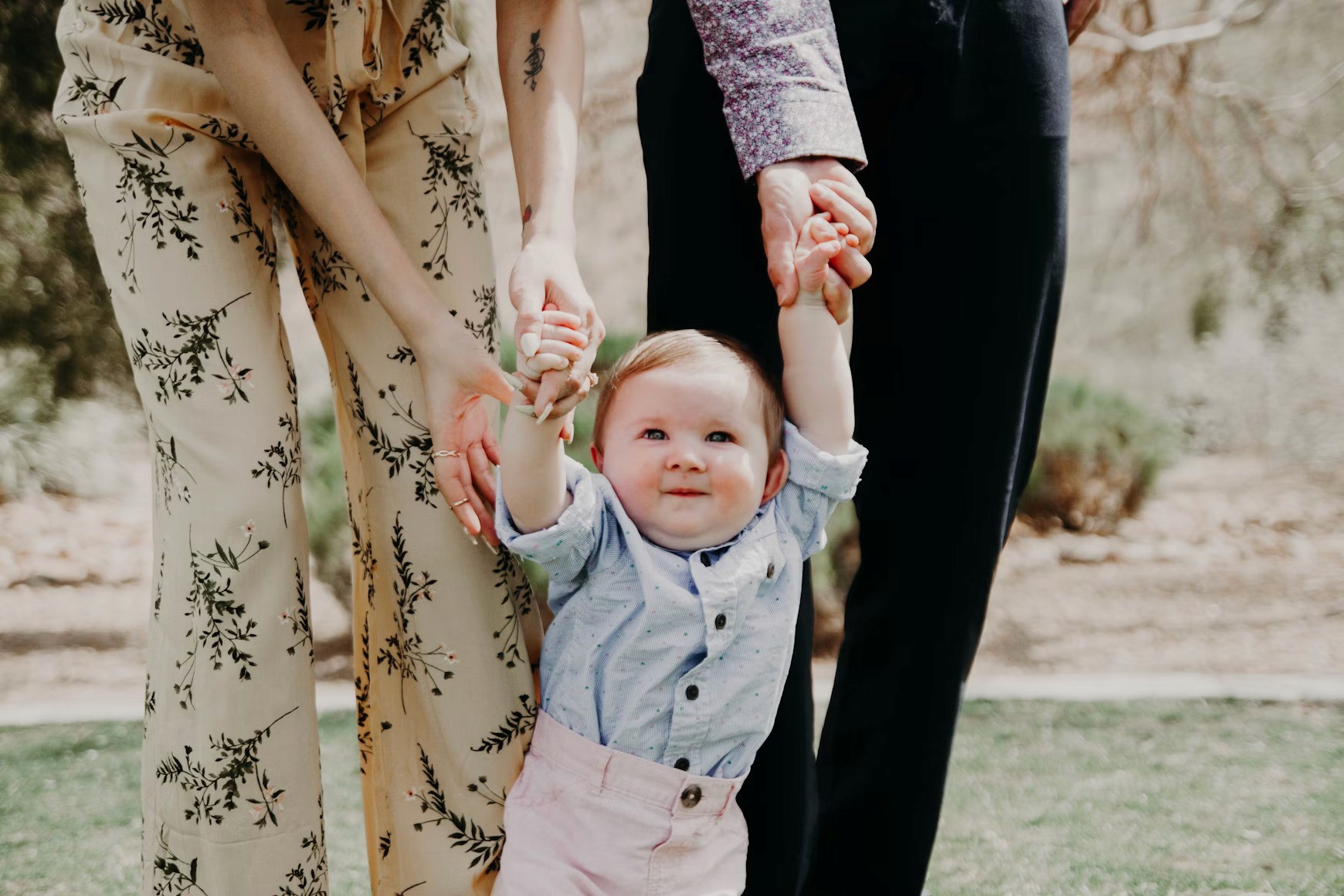 Baby holding hands with parents, smiling outdoors in sunny park.