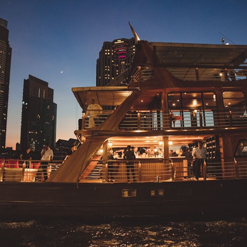 Un vapor cu mai multe punți plin cu oameni care iau cina și socializează, ancorat cu un fundal urban la amurg.