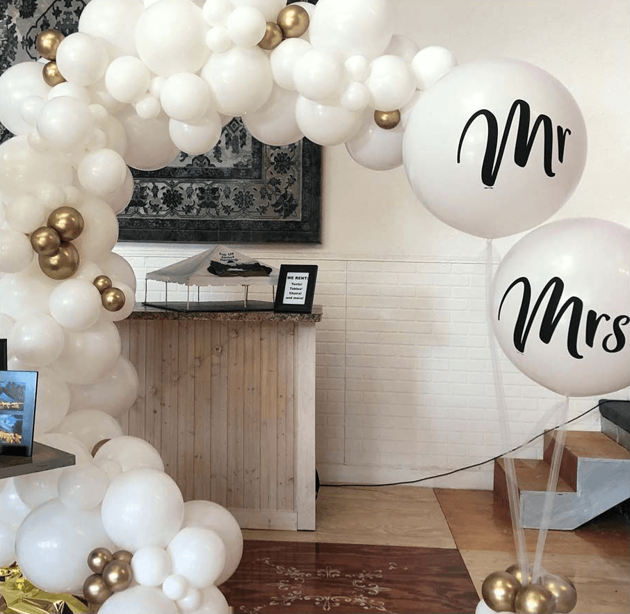 An elegant balloon arch made of white and gold balloons decorates the entrance of a venue, featuring a large "Mr" and "Mrs" balloon, creating a festive and welcoming atmosphere for a wedding celebration.