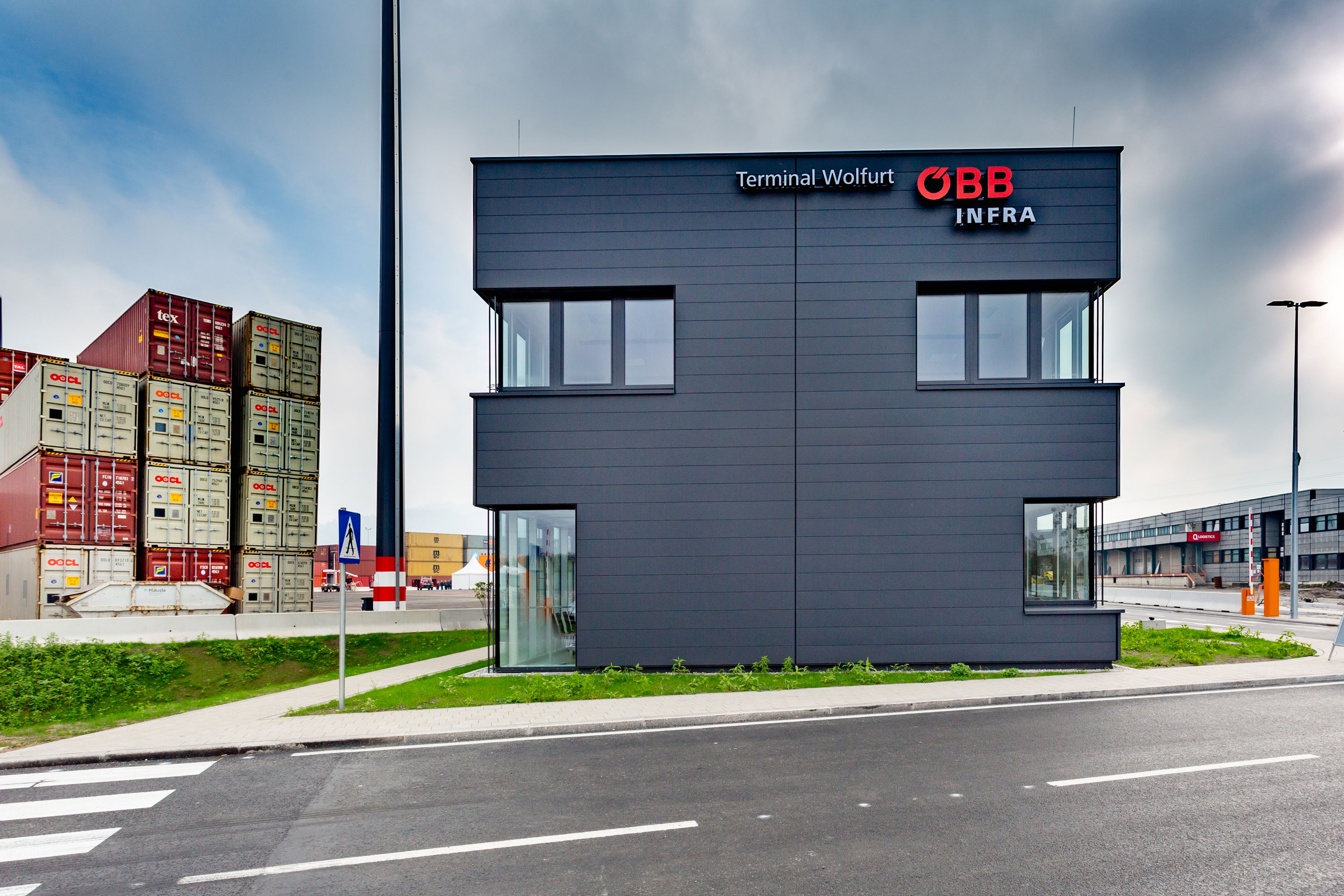 Dunkelgraues rechteckiges Gebäude mit großen Fenstern, Schriftzug 'Terminal Wolfurt ÖBB Infra' an der Fassade, gestapelte Container im Hintergrund.