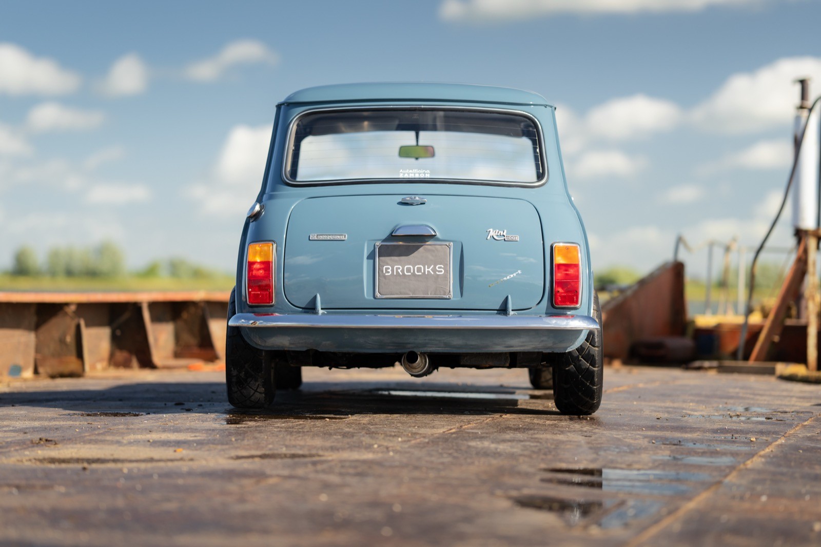 A small blue car parked on a gravel road, surrounded by open fields and a clear sky.