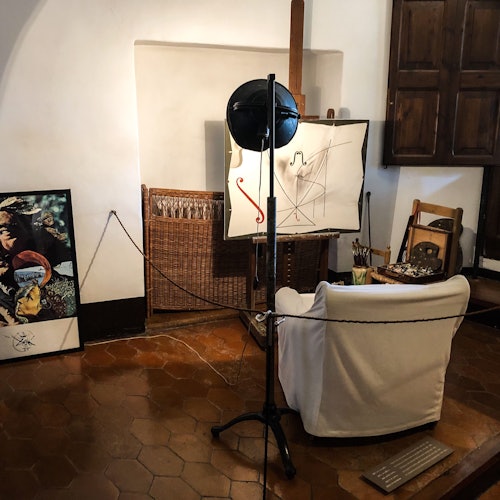 Art studio with a white chair, easel with abstract painting, overhead light, framed picture on the floor, and wooden furniture.