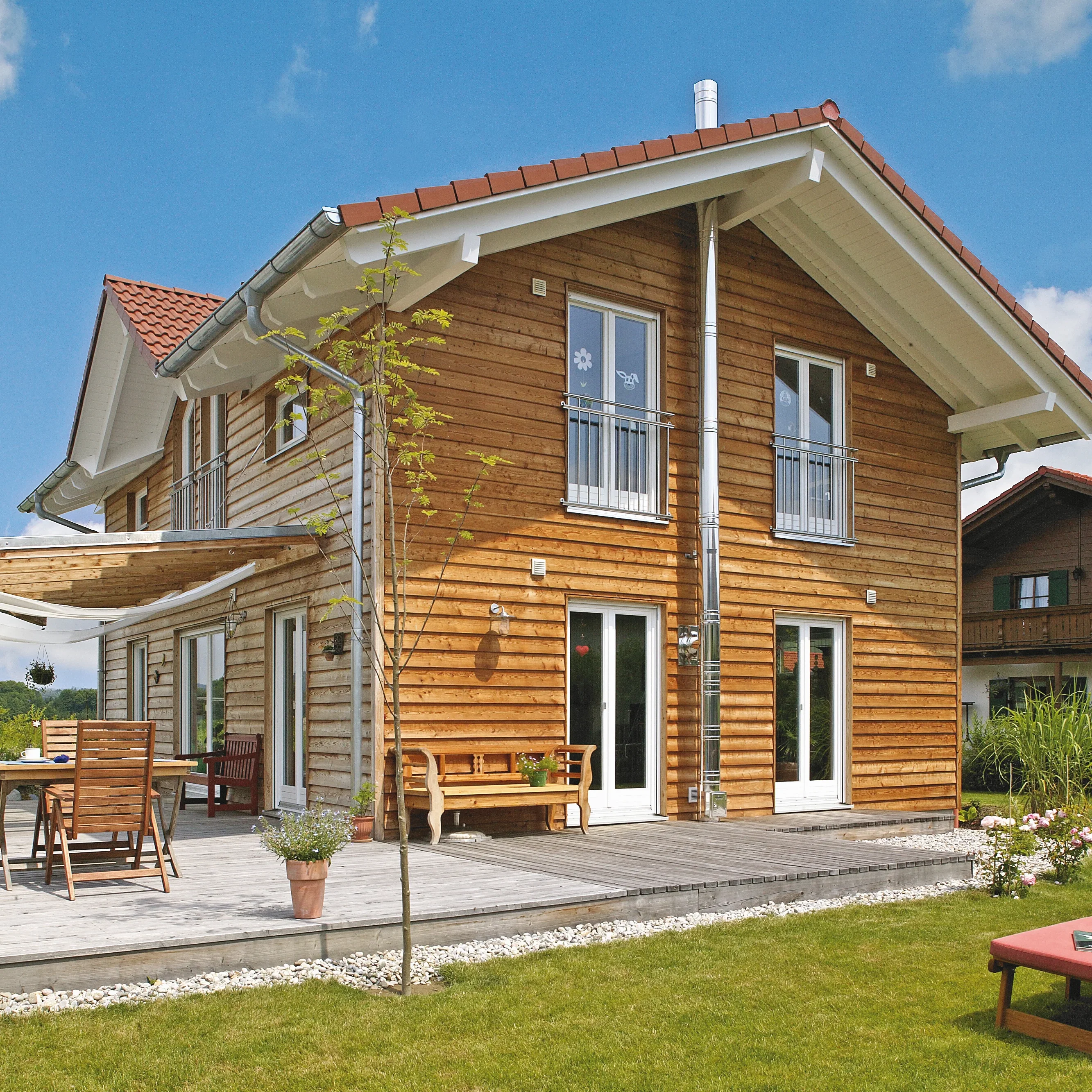 Geräumiges Haus mit Holzfassade und rotem Ziegeldach, umliegender Holzterrasse mit Gartenmöbeln & Rasenfläche.