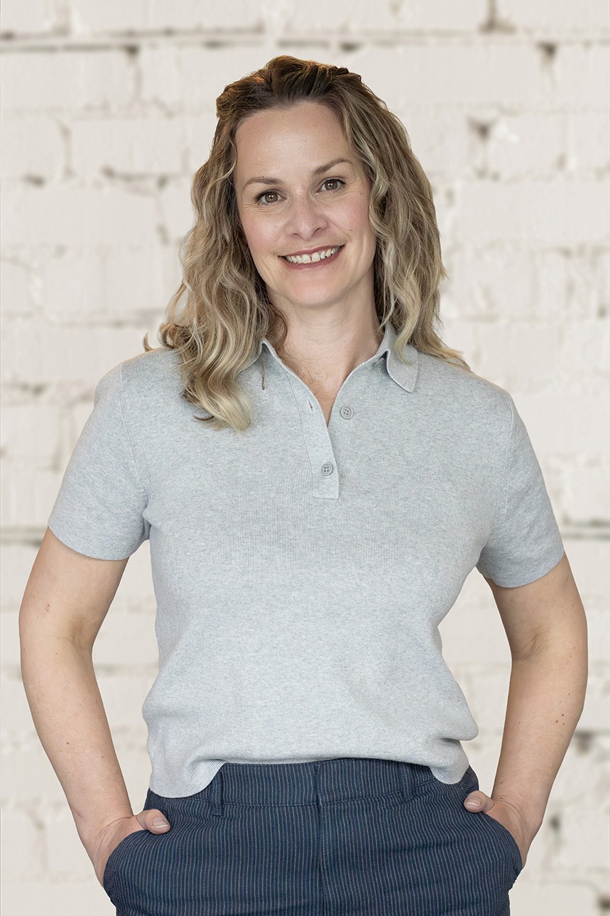 Image of Smiling woman with curly blond hair wearing a grey shirt, standing in front of a brick wall.