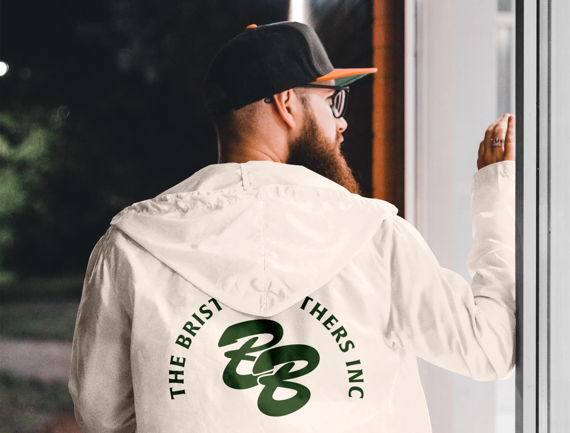 BB monogram printed on the back of a cream rain jacket being worn by a man with a beard wearing a flat brimmed hat.