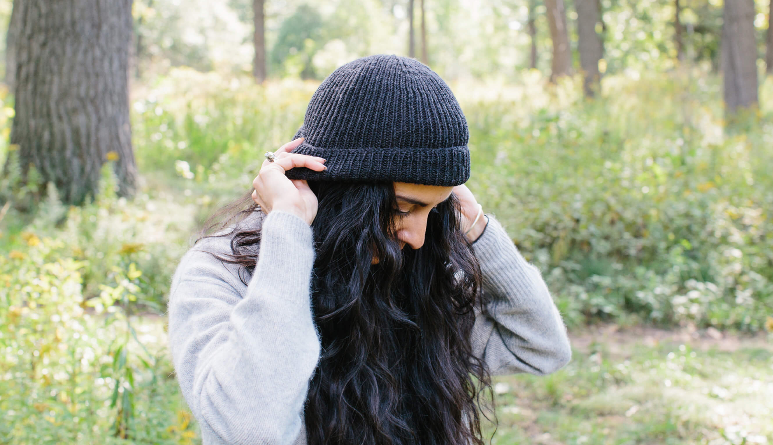 A women putting a dark grey tuque on her head, surrounded by forest.