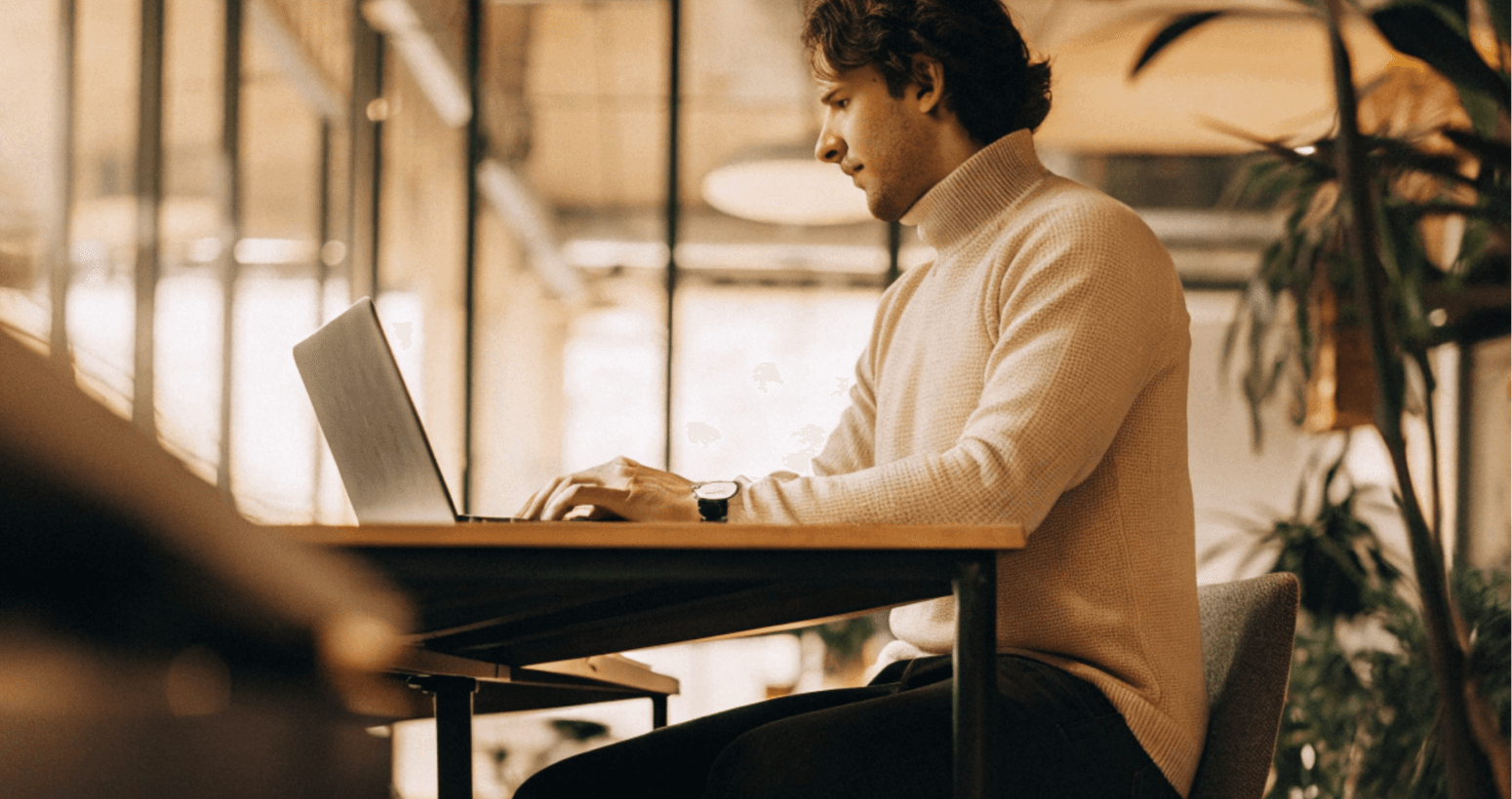 Man working on laptop at a table, focused on his work. Wearing a tan turtleneck in a modern office.