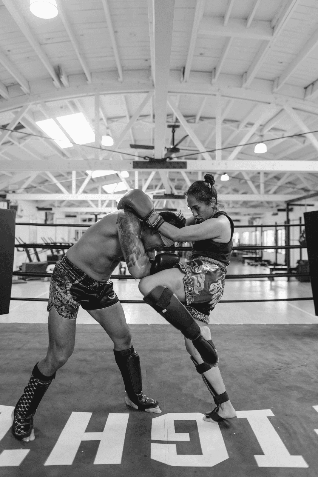 A female Muay Thai fighter delivering a knee strike while clinching an opponent inside a boxing ring during training.