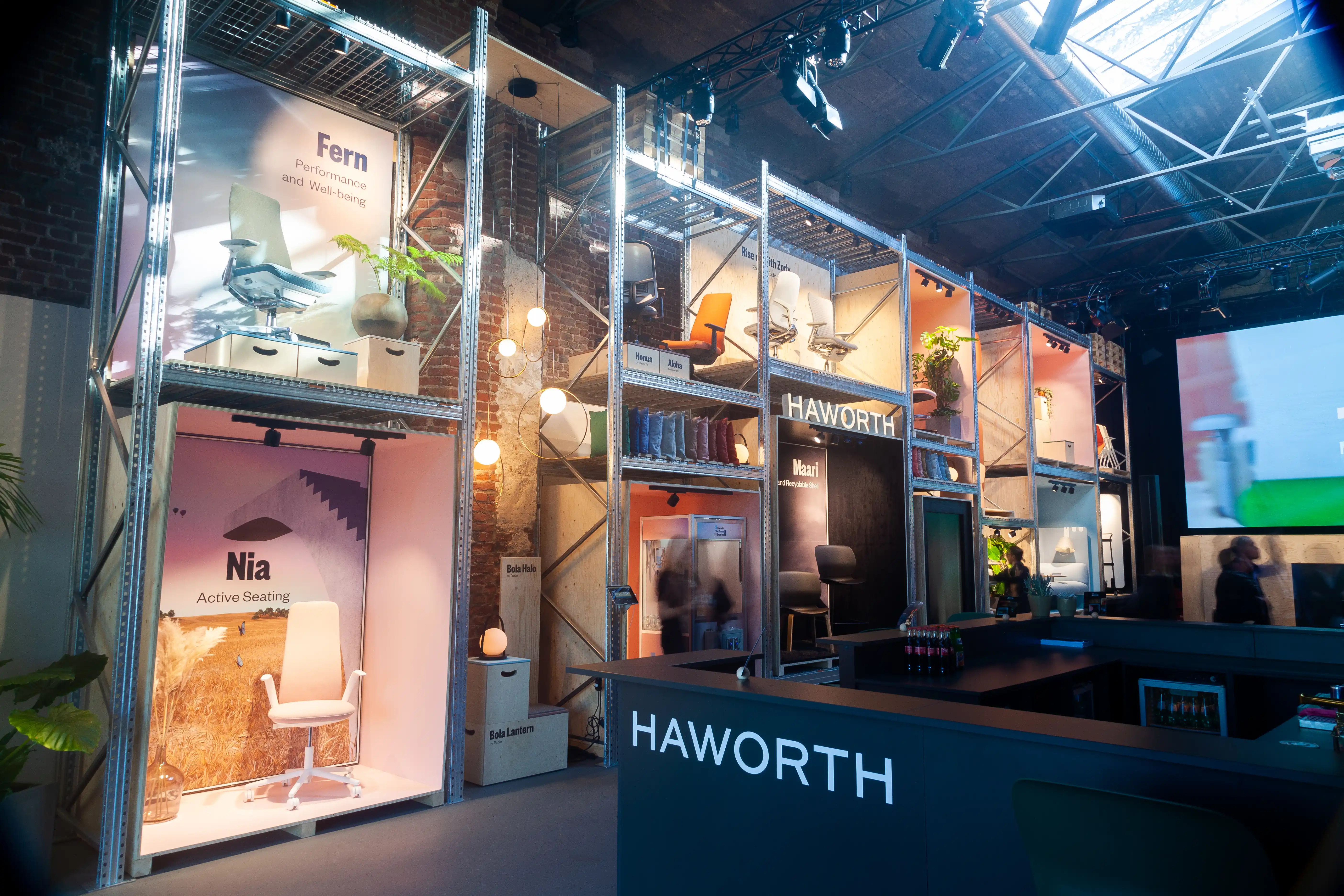 Haworth trade fair booth at ORGATEC with illuminated product boxes in scaffolding racks and a black bar counter in front.