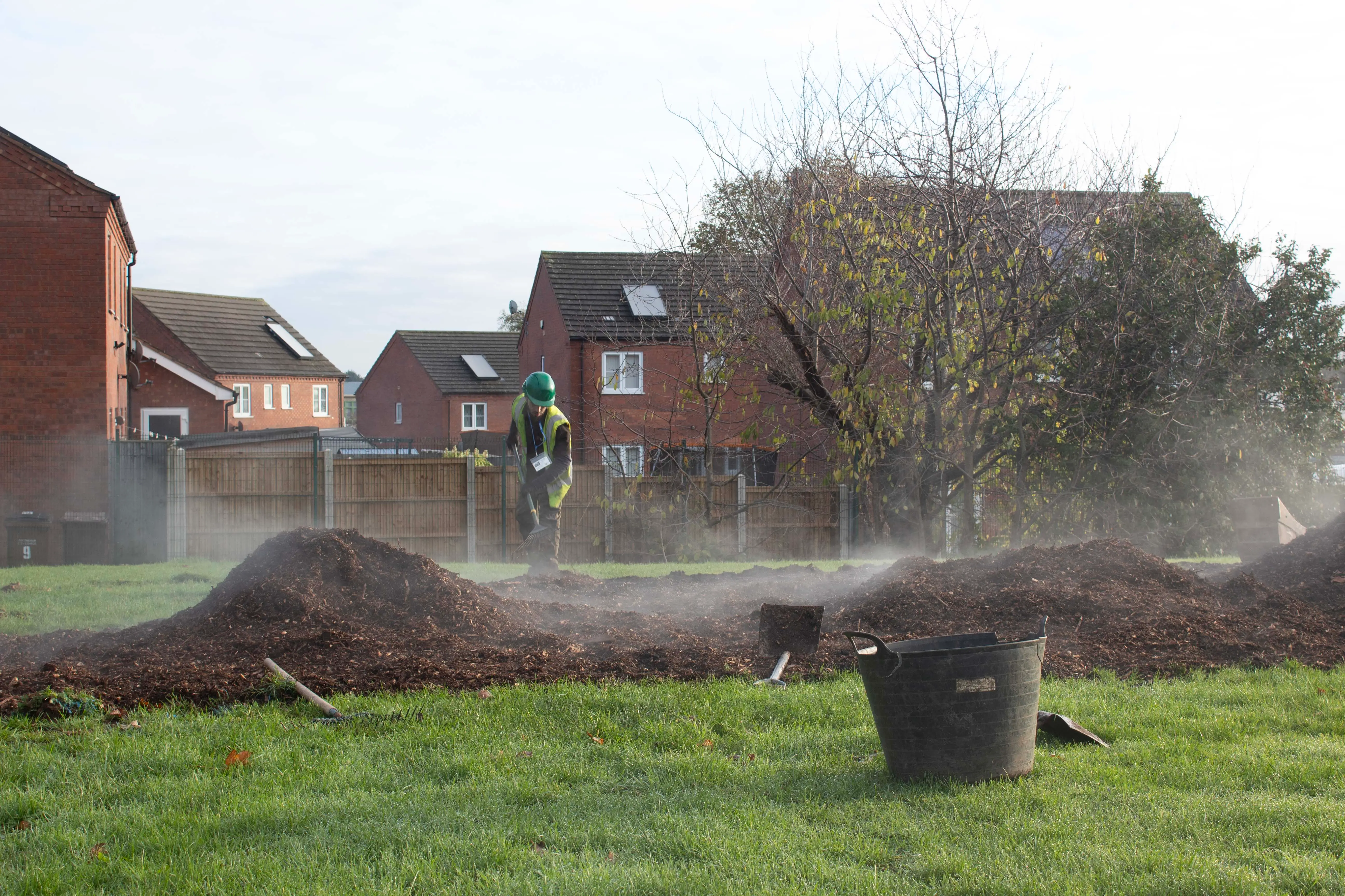 A grassy area with mounds of dirt and a few houses in the background, surrounded by mist or smoke.