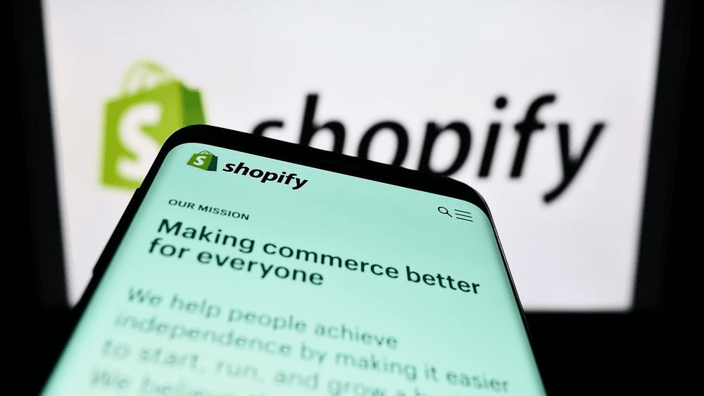 Smartphone displaying the Shopify website with the message “Making commerce better for everyone” in front of a blurred Shopify logo background.