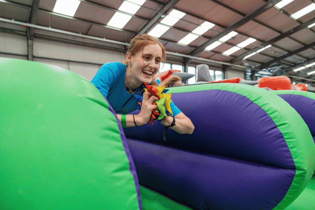 Outdoor team-building event with participants wearing matching blue shirts, taking part in inflatable obstacle courses and games, smiling and cheering, with the words 'ENTER A TEAM' in large bold white and green text across the image.