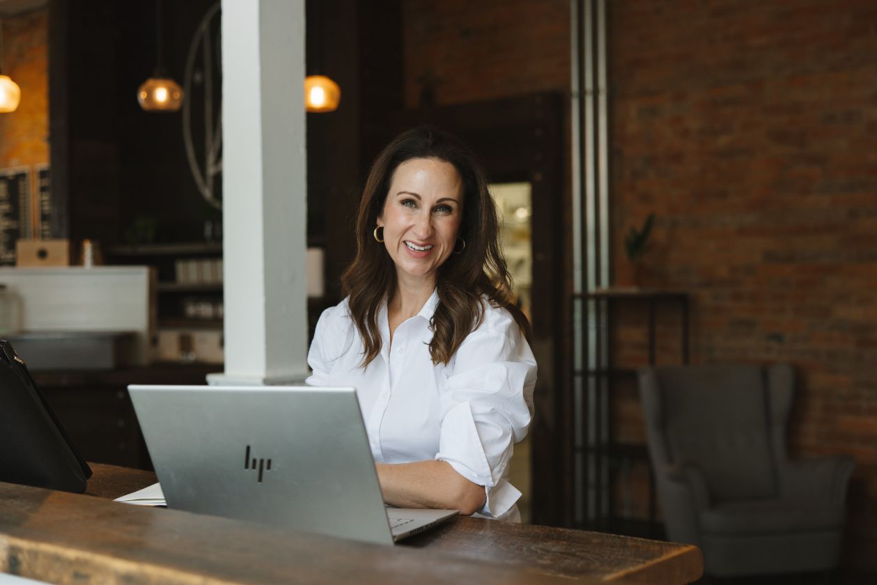 A smiling Stacy sits at a desk with a laptop, surrounded by a cozy workspace with wooden accents.