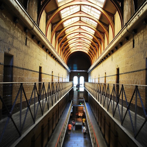 Interior corridor of a historical building with high arched ceiling, metal railings, and a symmetrical layout, leading to a bright window.