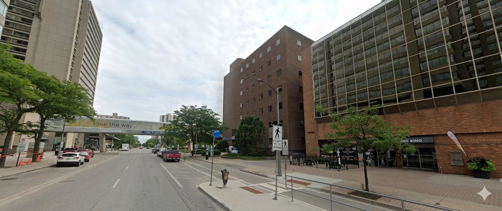 Street view of a city with mid-rise brick and glass buildings lining both sides of a one-way road. A skywalk with the words “Love the way you live” connects two buildings above the street, and pedestrians are seated outside a ground-floor restaurant on the right.