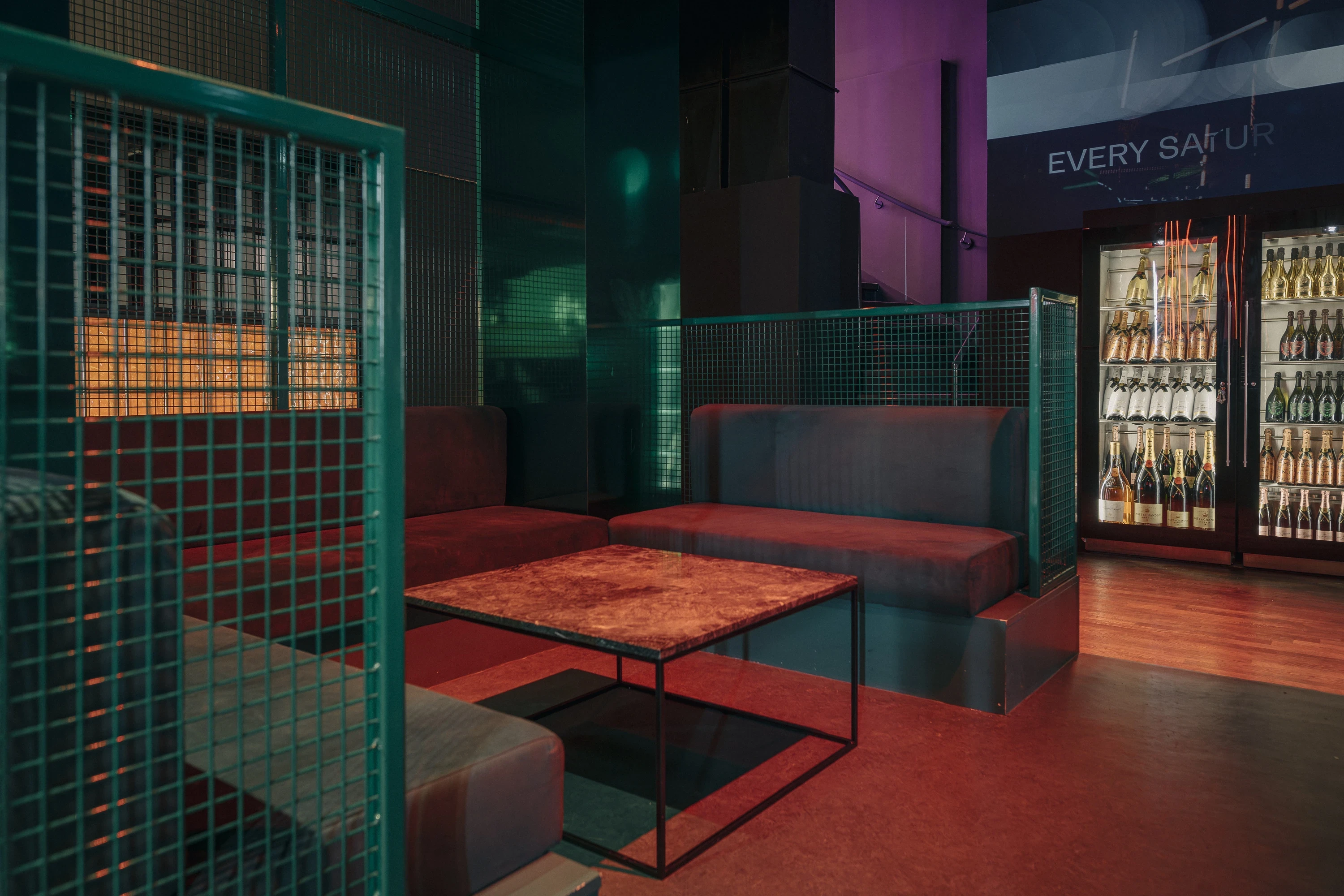 Lounge seating at NATE CLUB with dark red upholstered benches behind green metal mesh partitions, a red marble-look coffee table and illuminated drinks fridges.