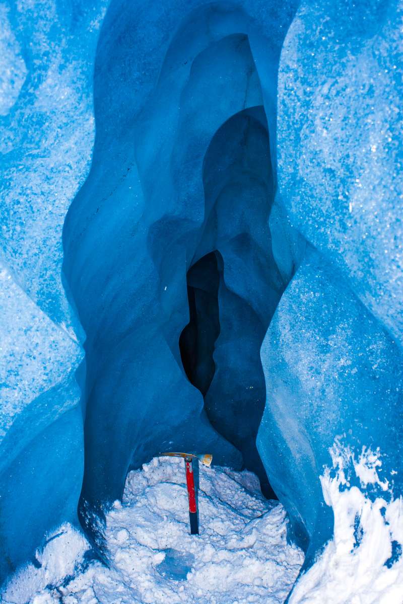 A person in a red and black jacket stands inside a deep blue ice cave with textured walls and white snow on the ground.