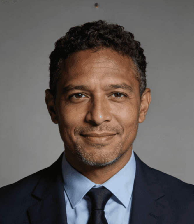 A professional portrait of a man in a suit and tie, smiling slightly against a neutral background.