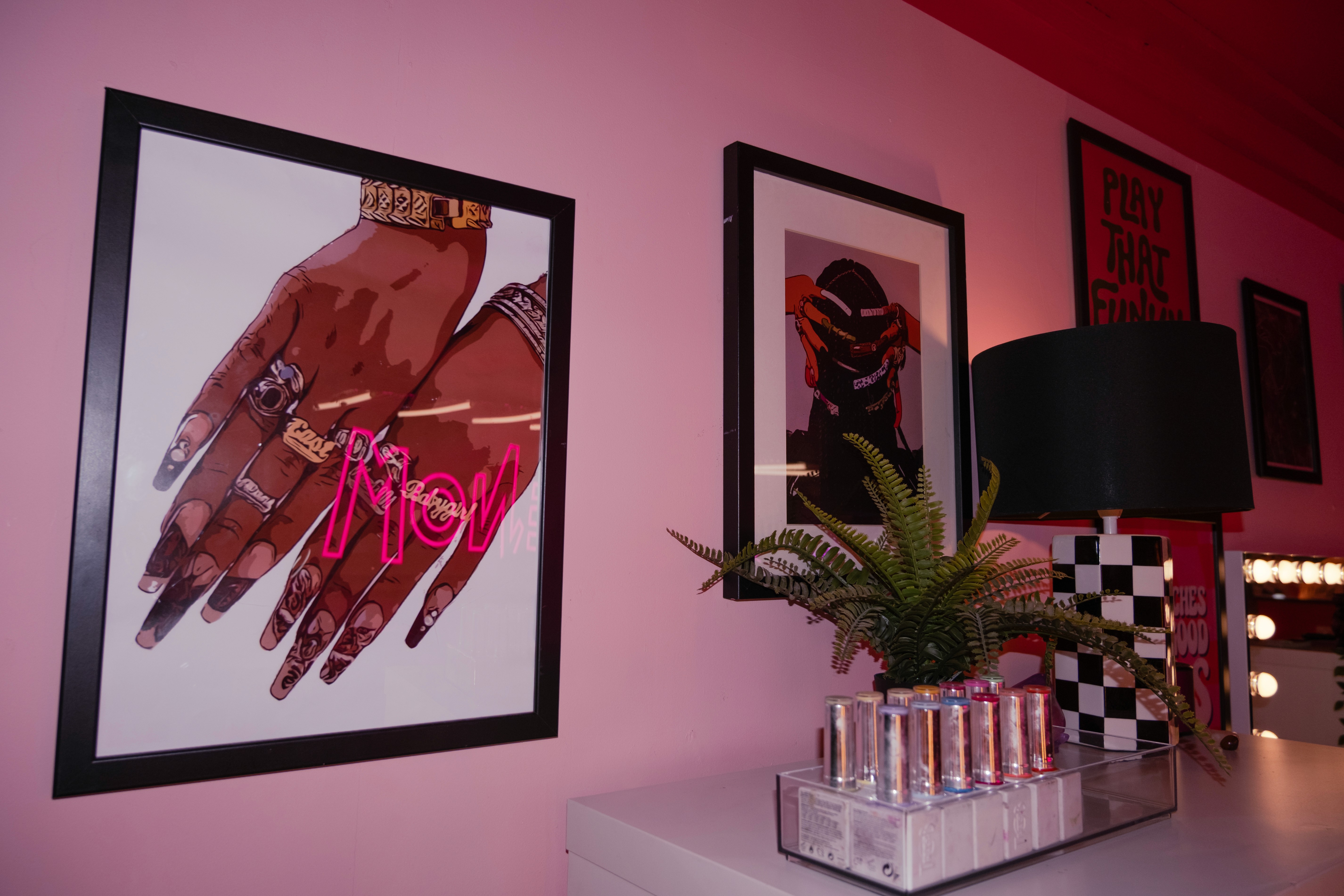 A close-up of framed nail-themed art on a pink wall, including a graphic of hands with long ornate nails and rings. A neon "VENOM" reflection is visible on the glass.