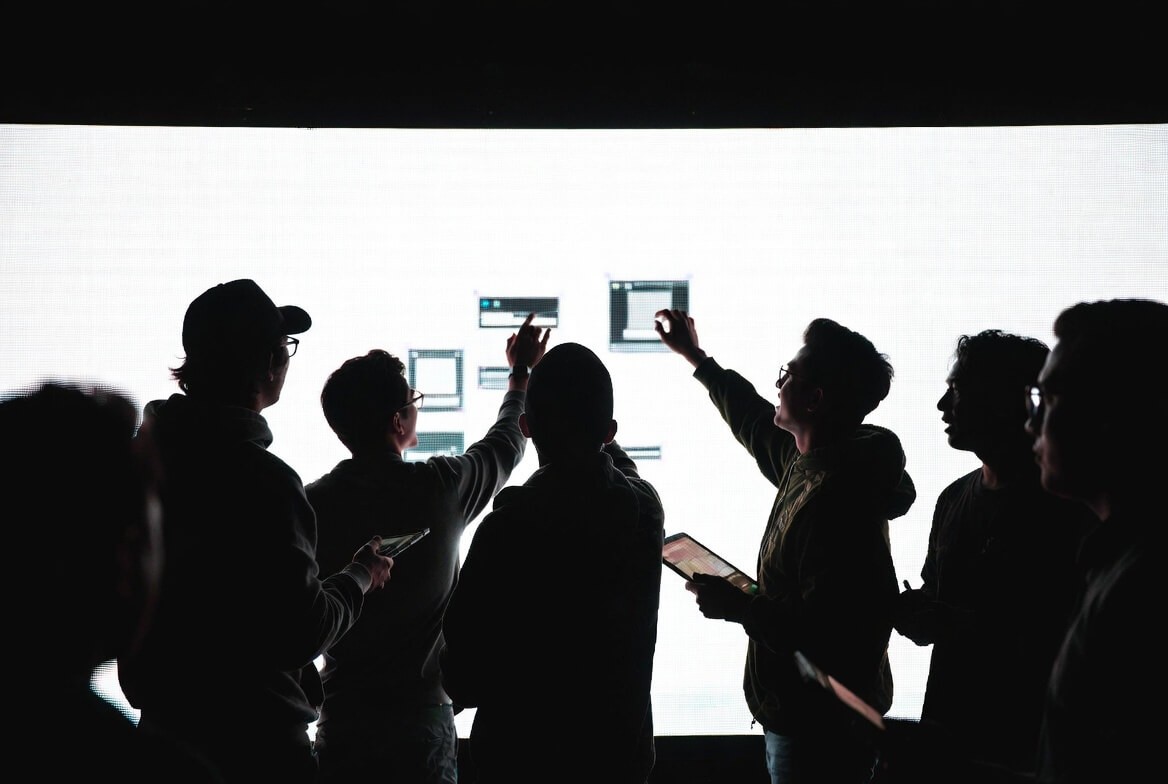 Team celebrating product launch. Silhouetted group on stage watching the big screen showing "Loading Complete" as one member presses deploy on a laptop. Moment of successful deployment and seamless transition. High-contrast silhouette style.