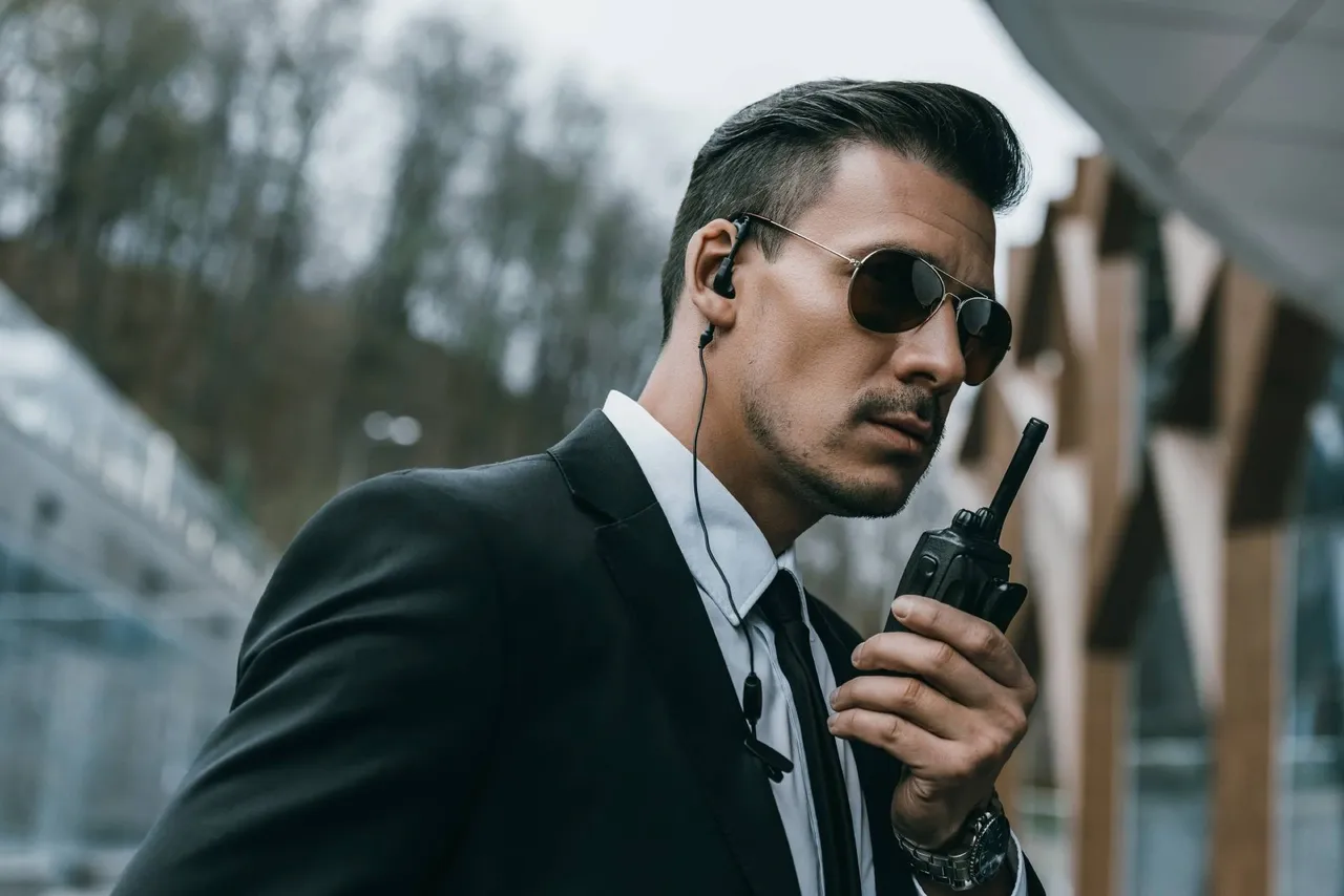 A serious security guard wearing formalwear and sunglasses talking on a walkie talkie.