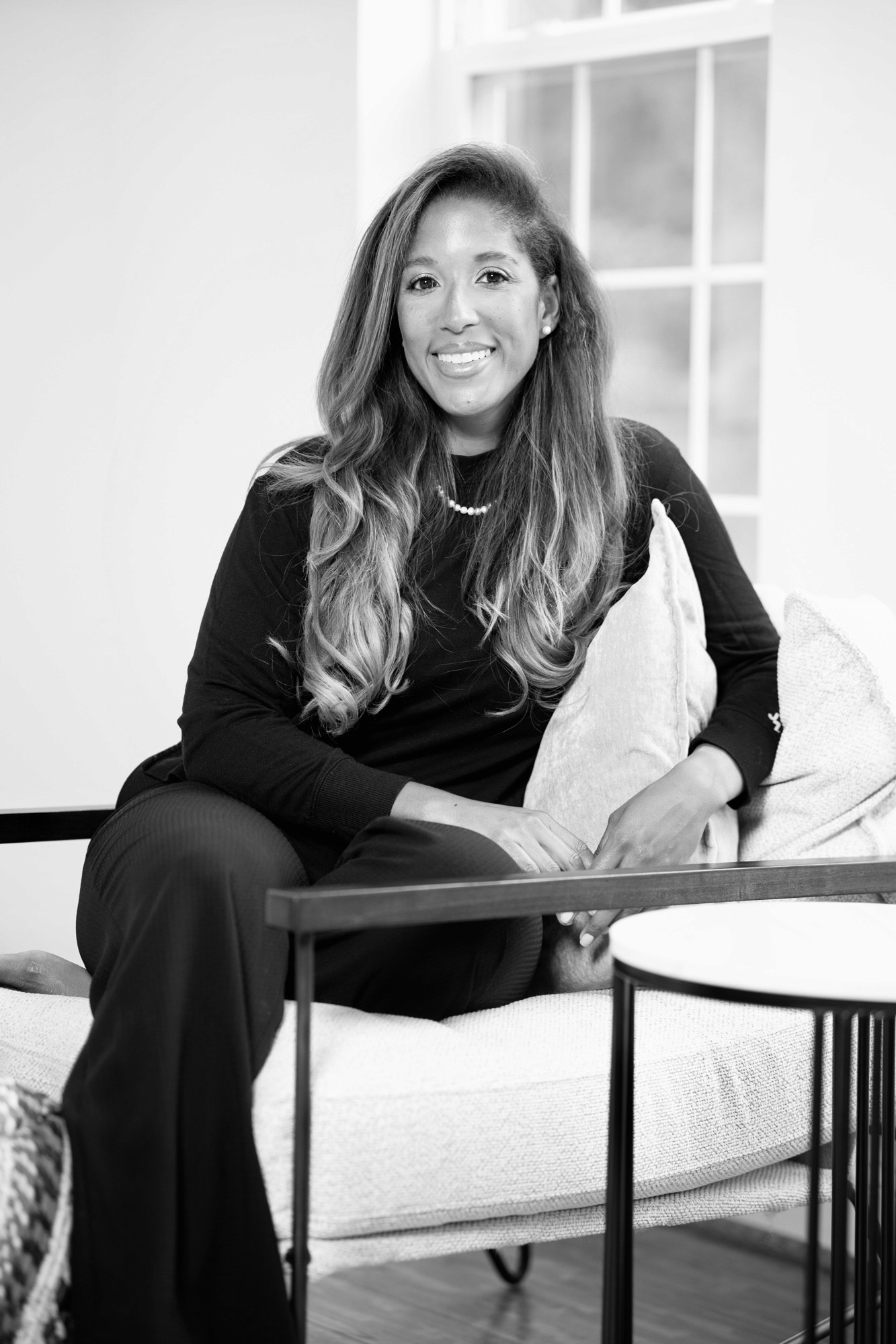 A smiling man with curly hair is sitting at a desk, looking at the camera, with a laptop and a lamp in the background.