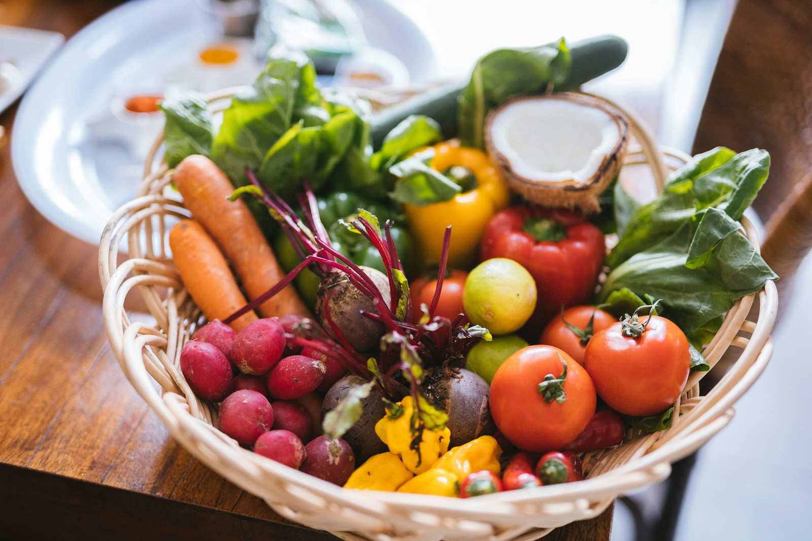 A woven basket filled with fresh vegetables including tomatoes, carrots, beets, and leafy greens on a wooden table.