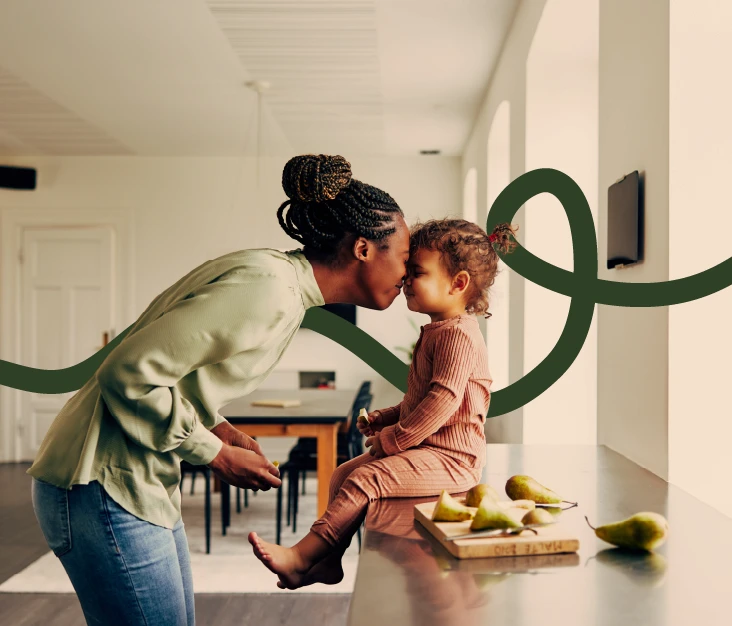 A Black mother leaning in for a nose kiss with her toddler sitting on a kitchen counter, surrounded by pears on a cutting board. A dark green curving line graphic from the Tafe brand overlays the image.