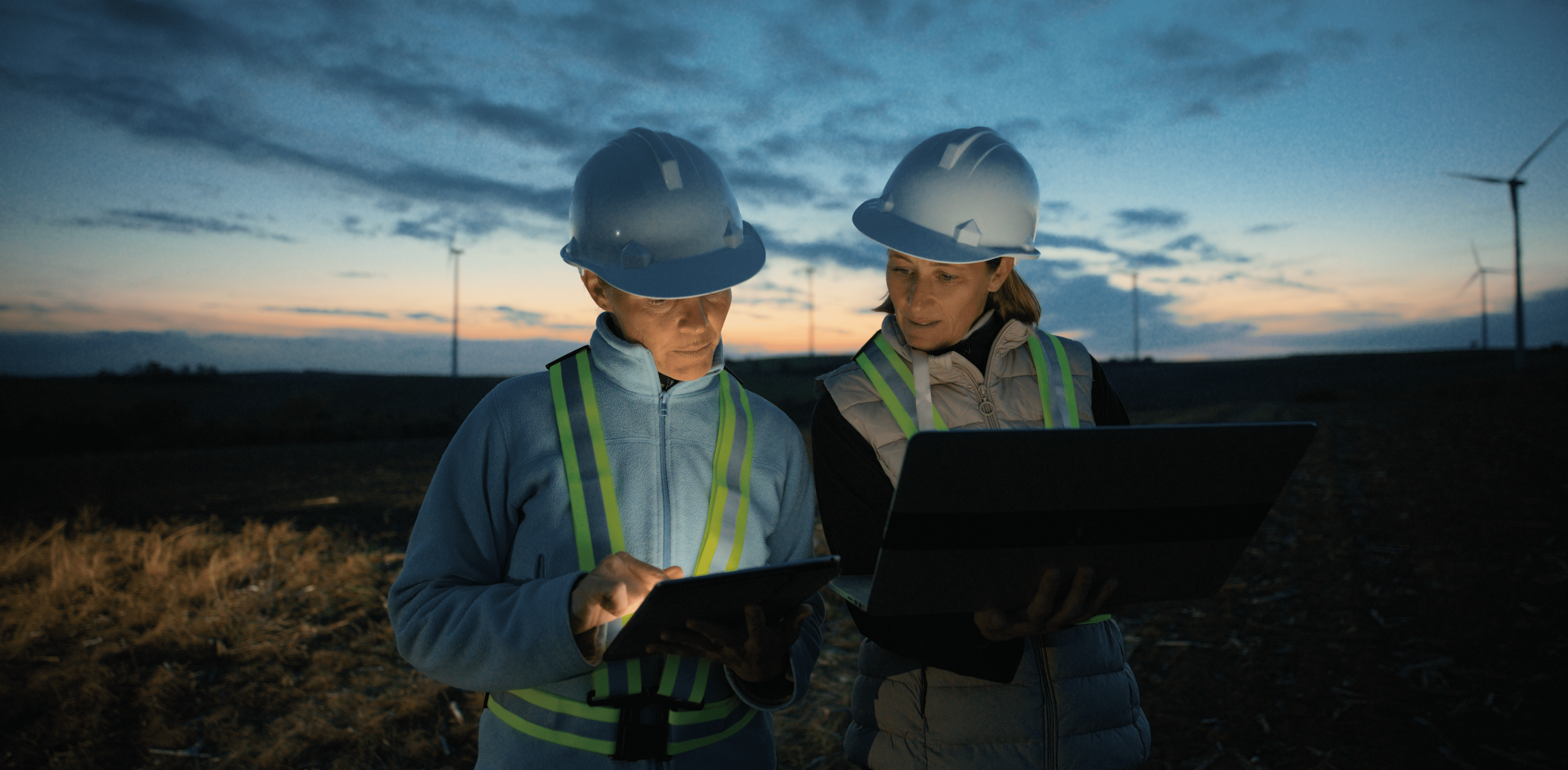 Two female engineers wearing white hard hats and high-visibility safety vests stand in a field at dusk, reviewing documents on tablets using the Voltquant app. Wind turbines are visible on the horizon against a cloudy sky, indicating they're working on a wind energy project.