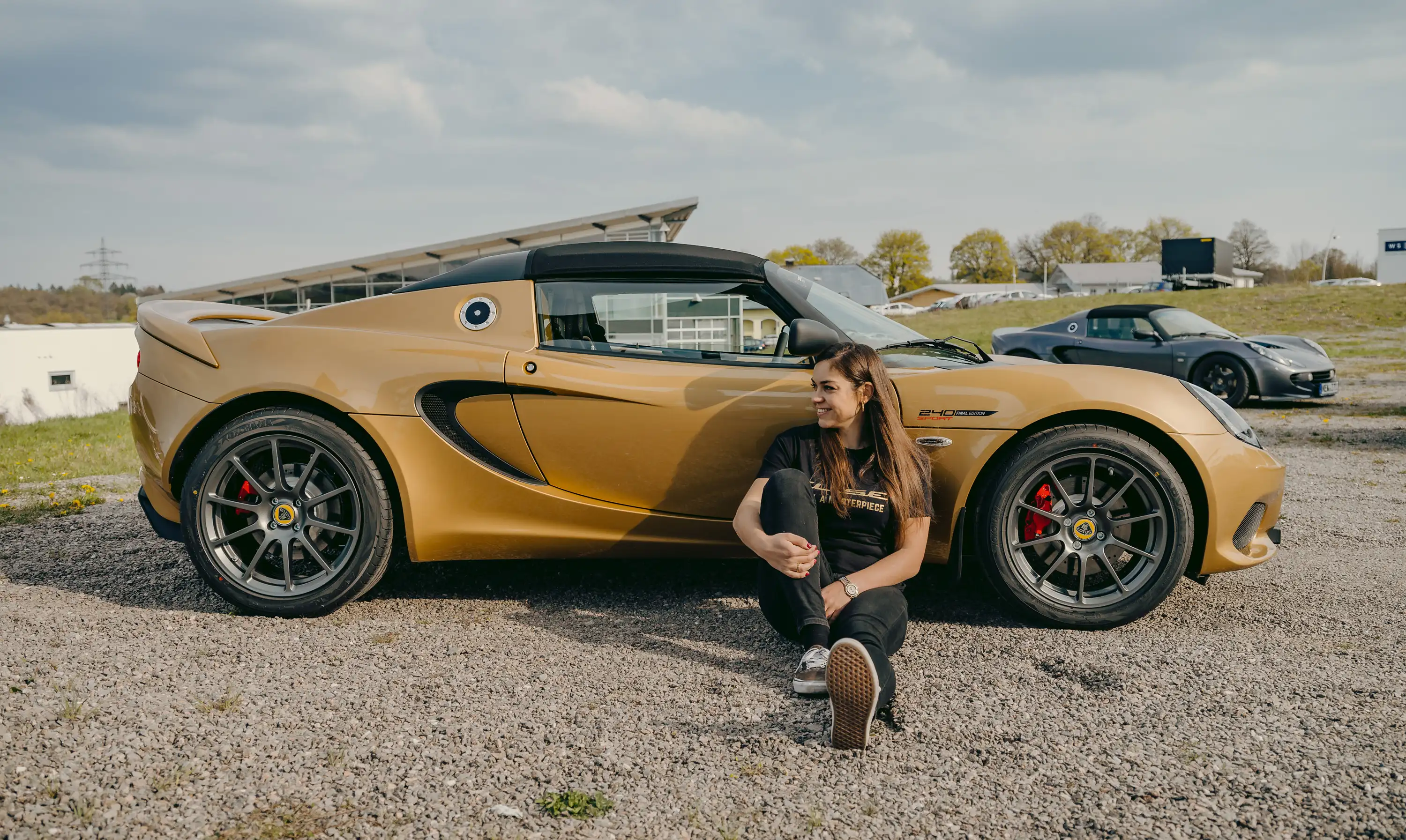 Elisa Artioli sitting on the ground, smiling next to a gold Lotus Elise 240 Final Edition parked on gravel, with another Lotus visible in the background.