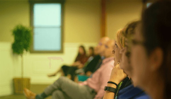 A small group of people seated in a room, listening attentively, with a woman in the foreground looking thoughtfully to the side, suggesting a meeting, seminar, or group session.